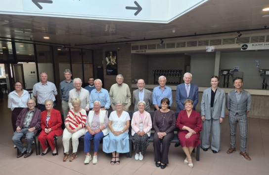 Groepsfoto van 20 mensen, zittend en staand in een hal met pijlen aan het plafond. (vergroot foto)
